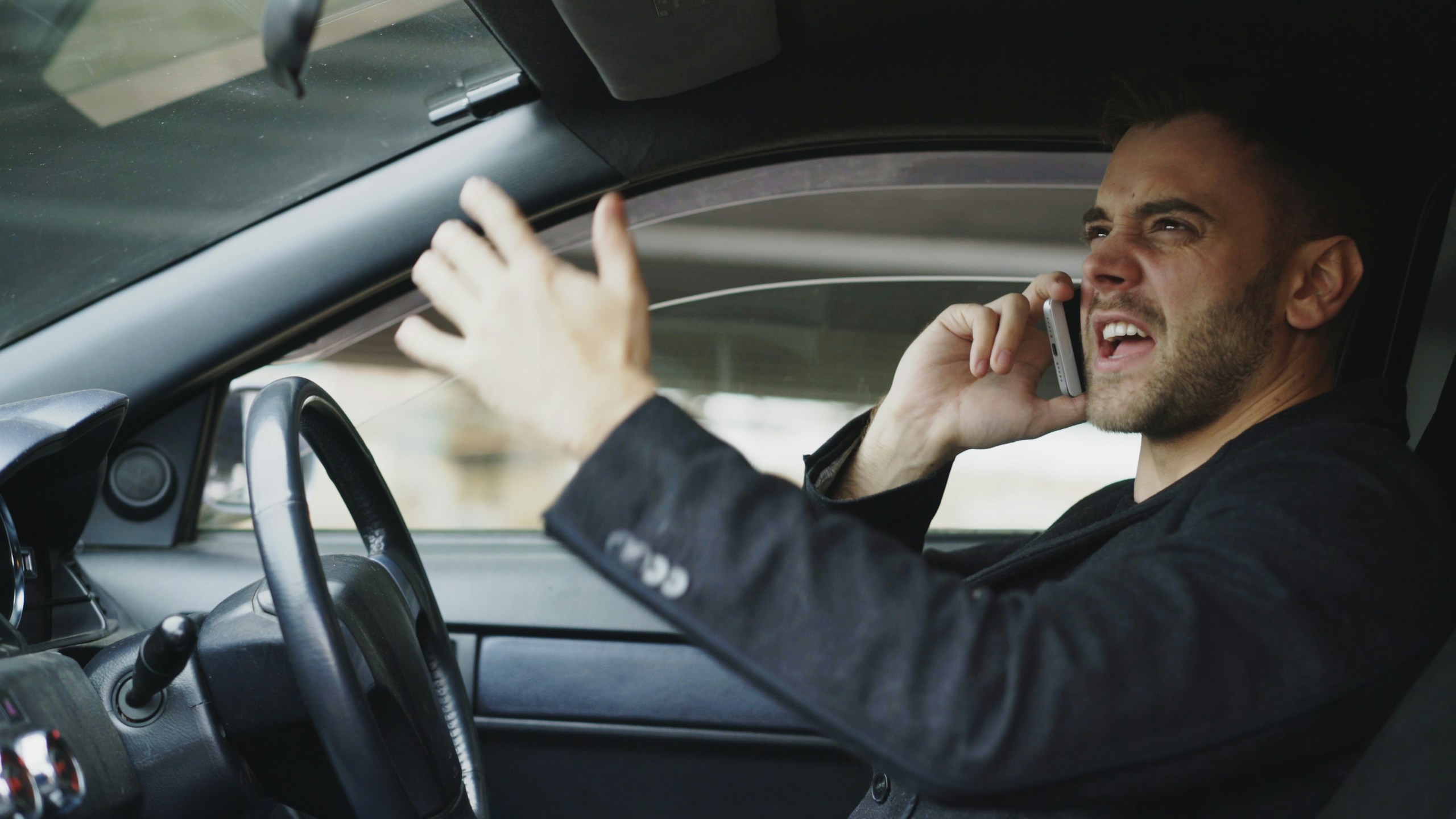 Stressed young businessman swearing and talking phone while sitting inside car outdoors