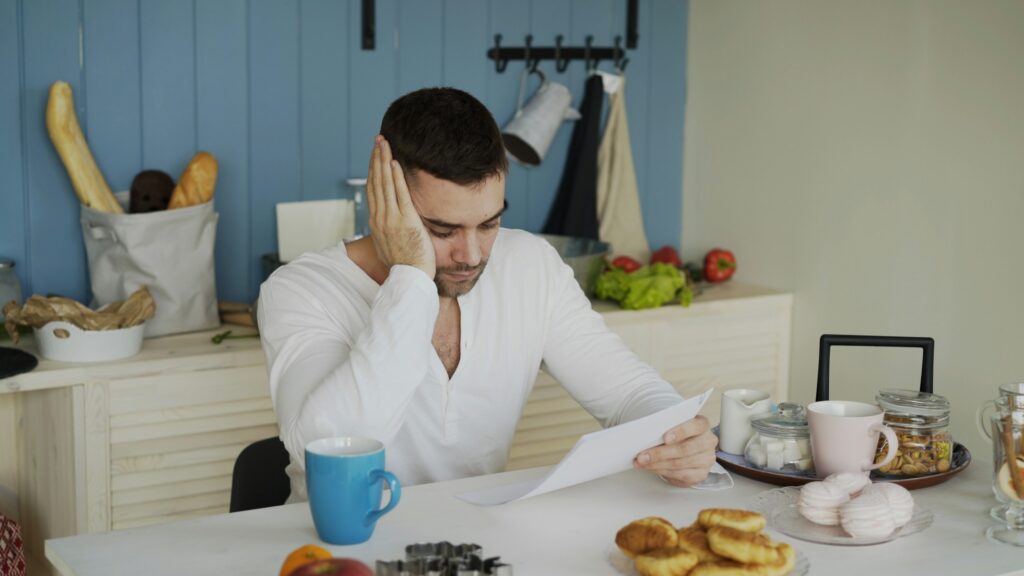 Upset young man reading letter with unpaid bill in the kitchen at home eraly morning