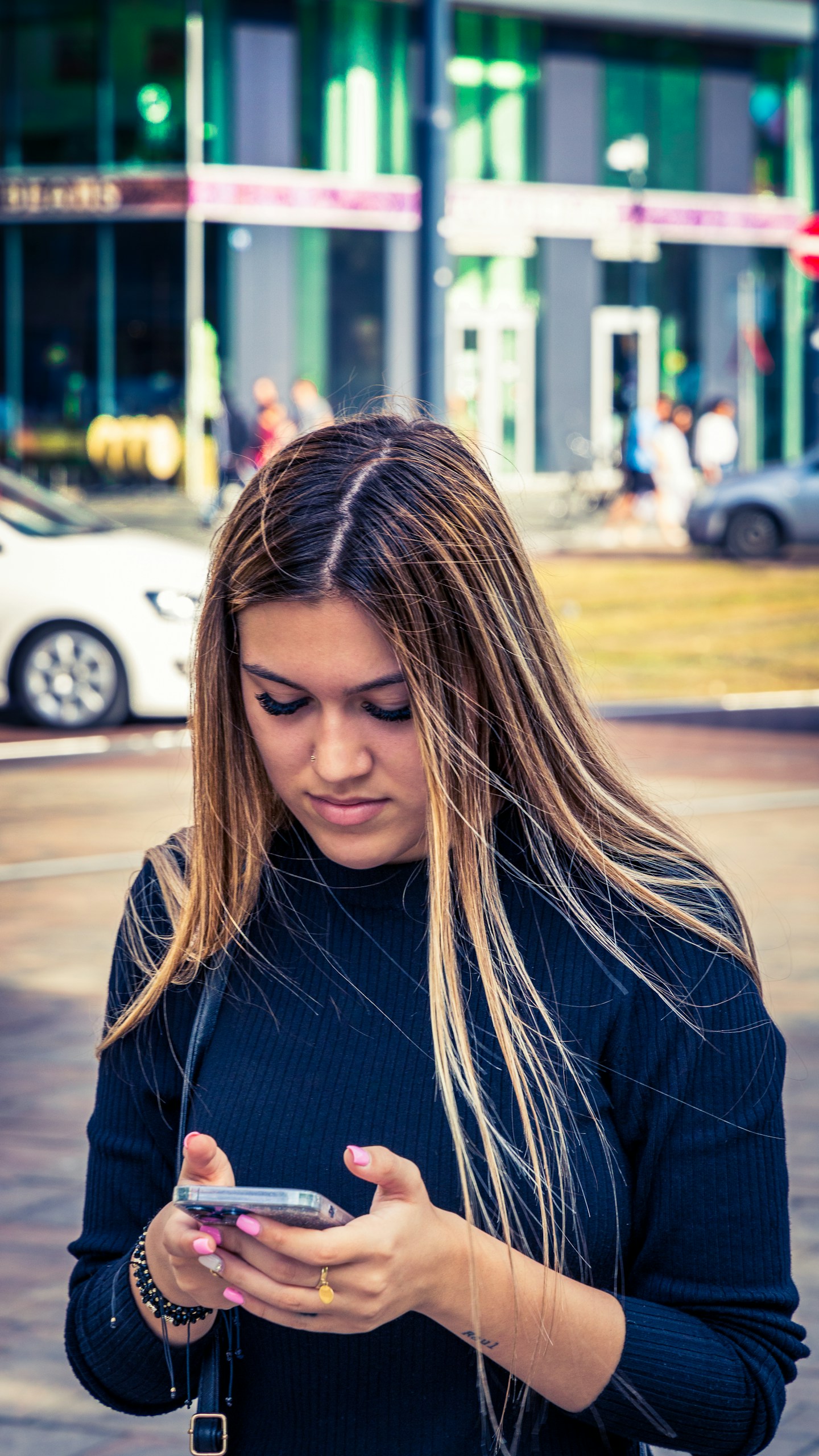 Young girl reading a text message on a sunny day in the city A young beautiful girl with blonde-brownish hair, dressed in a black hoodie, holding a phone with both hands, and looking at it while standing on a sunny day in the street. She is in the shade of a building at an intersection with a few cars passing by and people walking. There is a building with glass and metal in the background, indicating that she is in the center of a city. This photo shows a young woman standing on a sunny day in the city. She is wearing a black hoodie and holding a phone in both hands, reading a text message. T