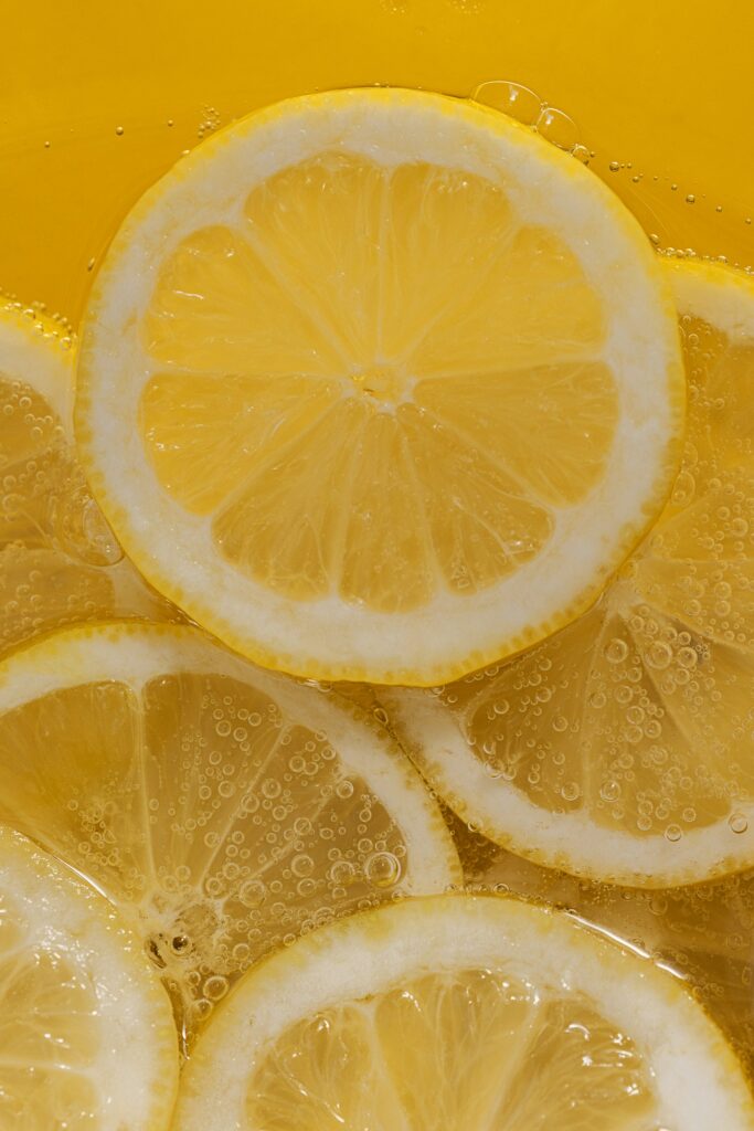 A close up of lemon slices in tonic water. The tonic water has created little bubbles on and around the lemon slices. The image is set against a yellow background.
