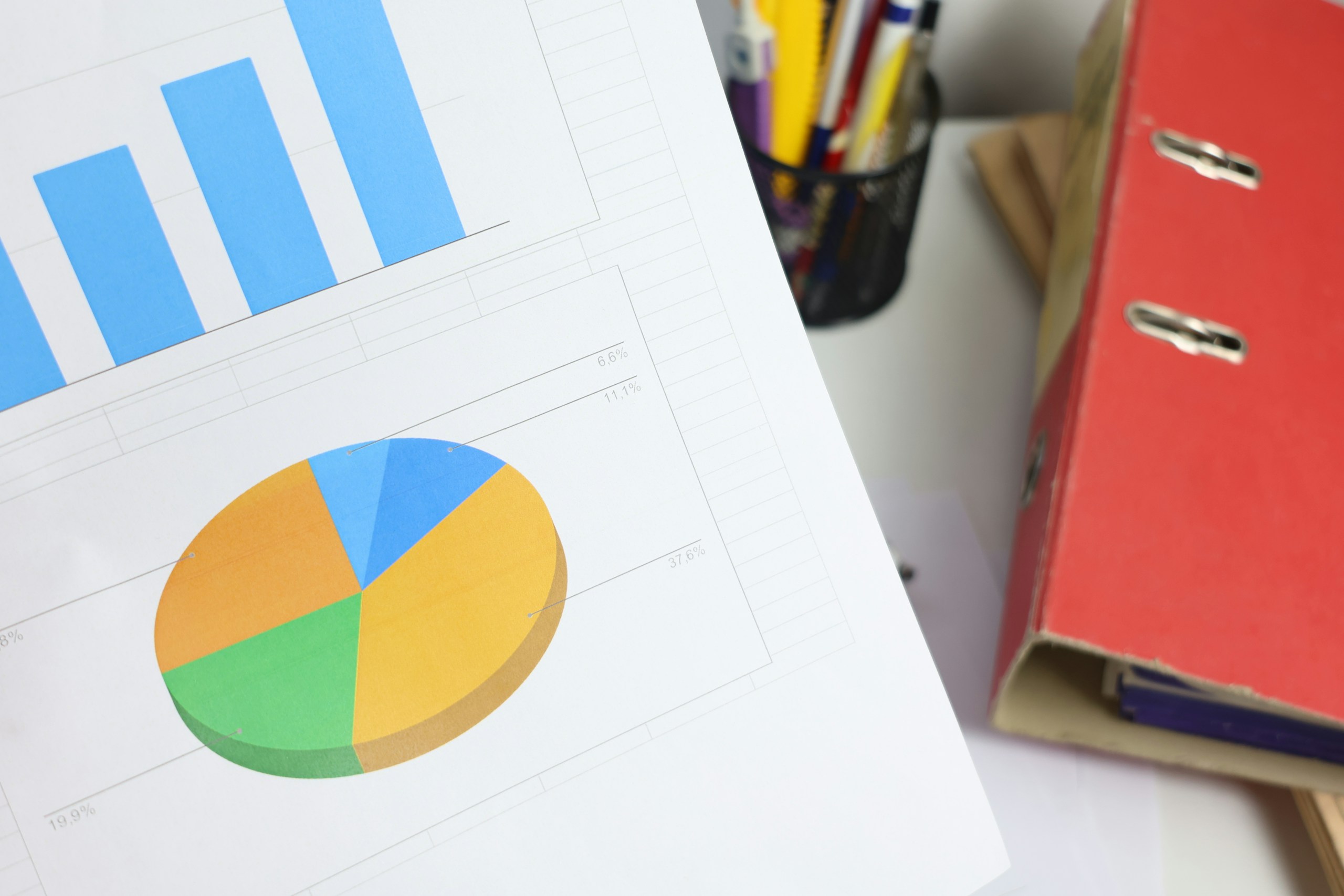Printed reports on a desk showing a blue bar chart and a multicolored pie chart, with a red binder nearby.