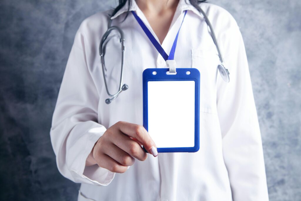 Medical professional in a white coat wearing a stethoscope around the neck and holding a blue ID badge on a lanyard with a blank white card.