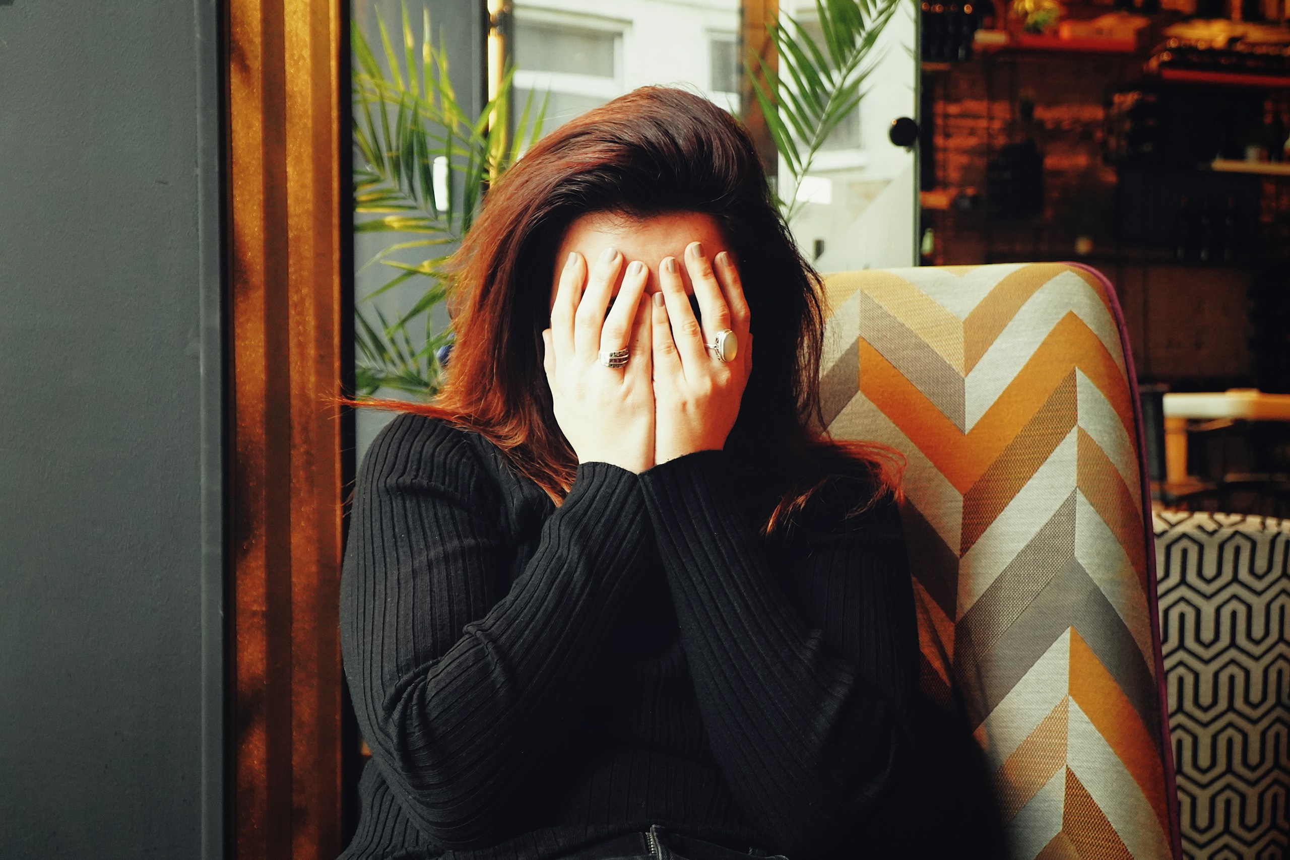 Person with hands covering their face, sitting on a patterned chair in a cozy indoor space.