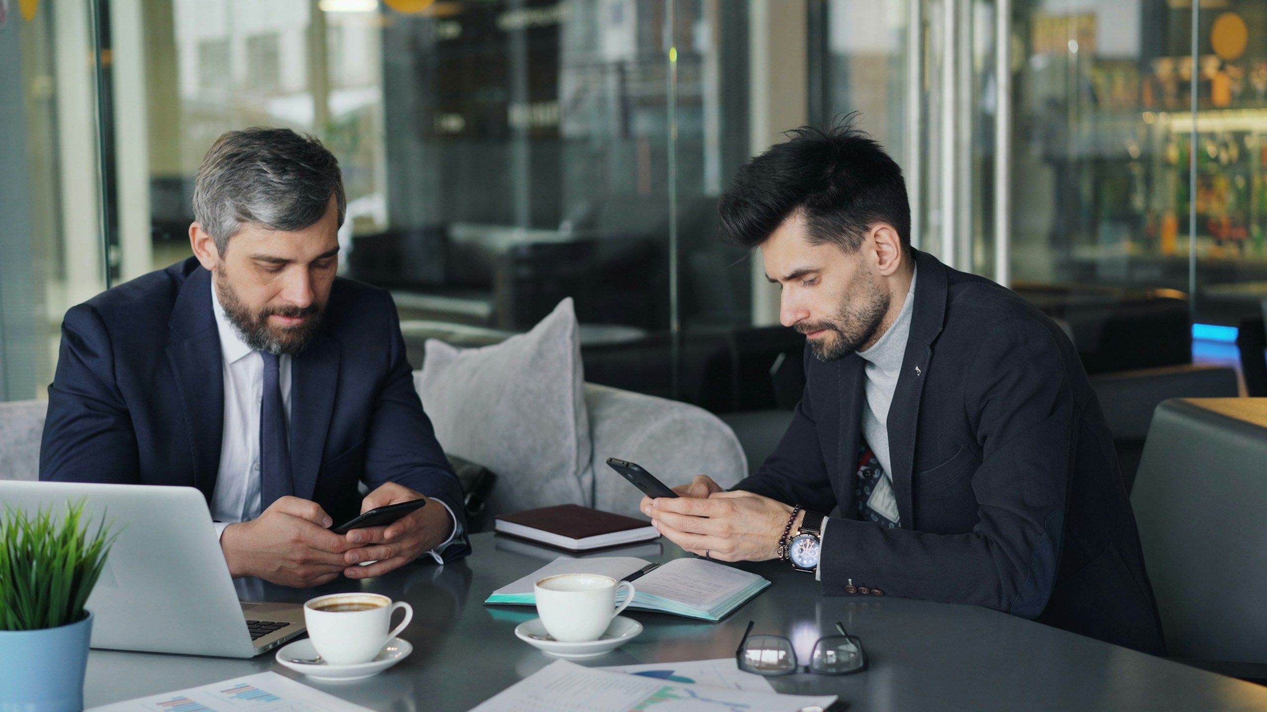 Two businessmen in suits sit at a glass-walled office, looking at their smartphones with a laptop and papers on the table, coffee cups nearby.