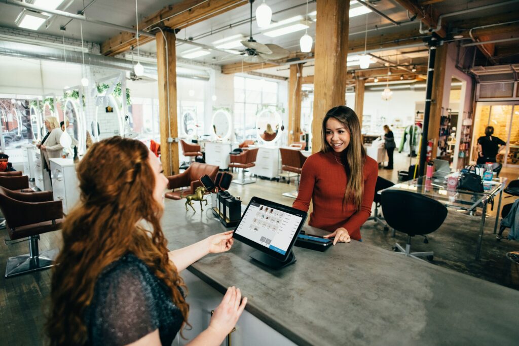 Hair salon interior with wooden pillars, a stylist and client at the reception counter looking at a tablet screen amidst styling chairs and mirrors in the background.