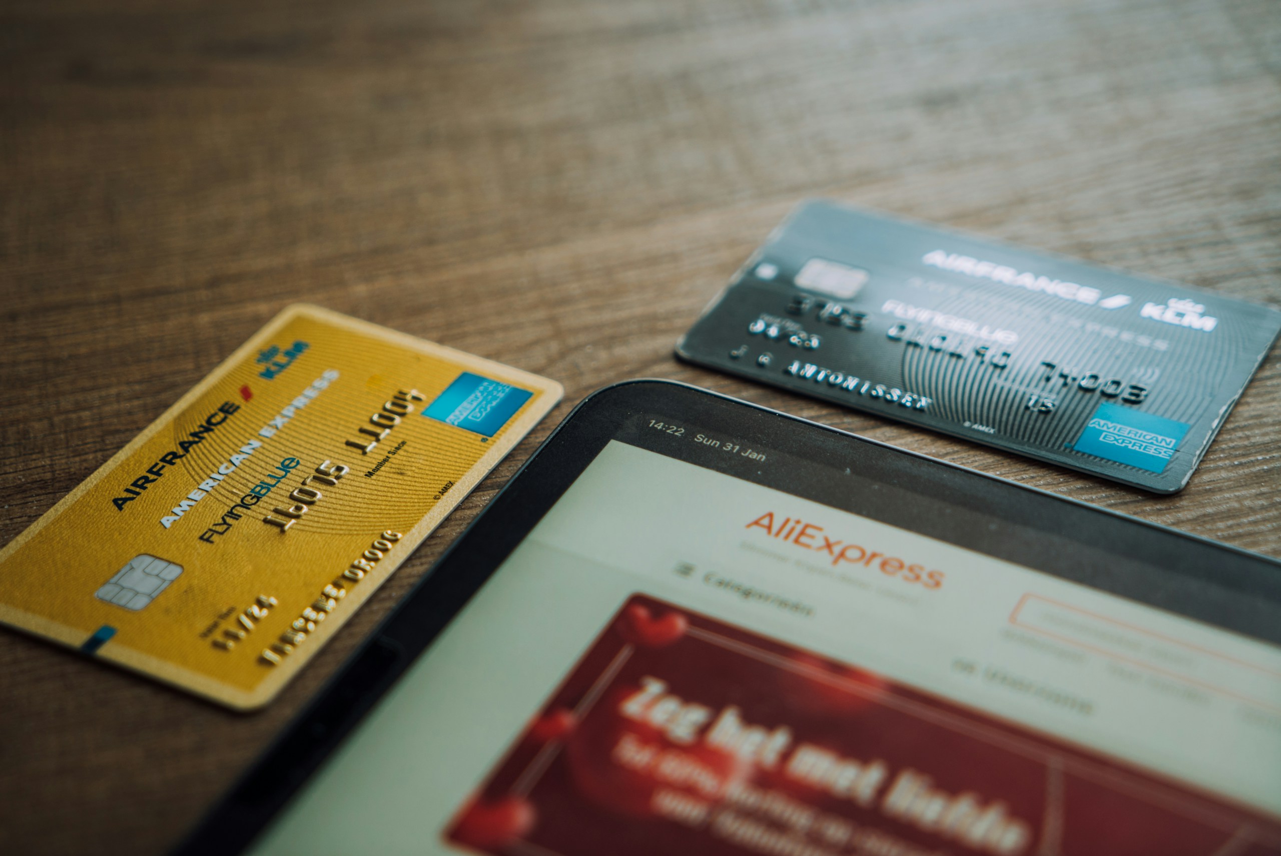 Credit cards resting on a wooden table beside a tablet showing a shopping site (AliExpress) titled page content blurred in the foreground.