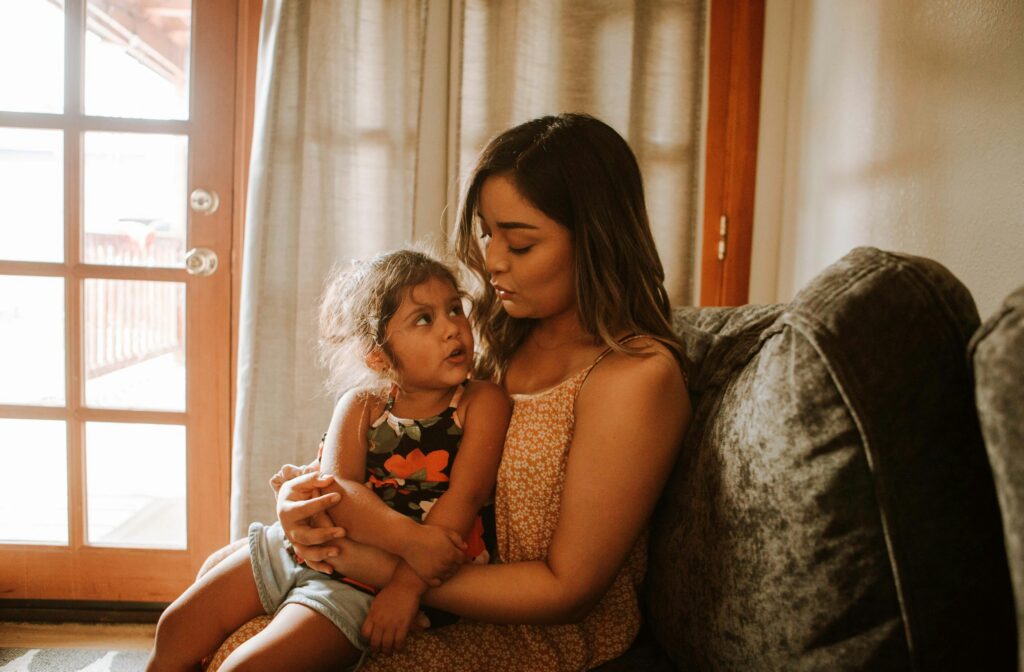 A woman sitting on a sofa holding a young child, both looking at each other warmly indoors near a glass door.