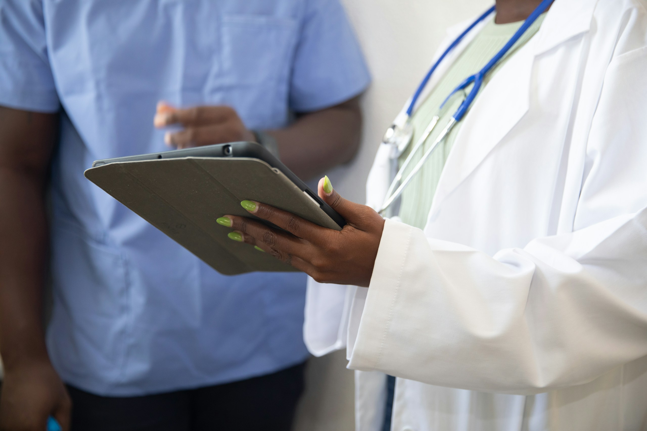 Two healthcare professionals—one in a white coat with a stethoscope and the other in blue scrubs—reviewing content on a tablet device.