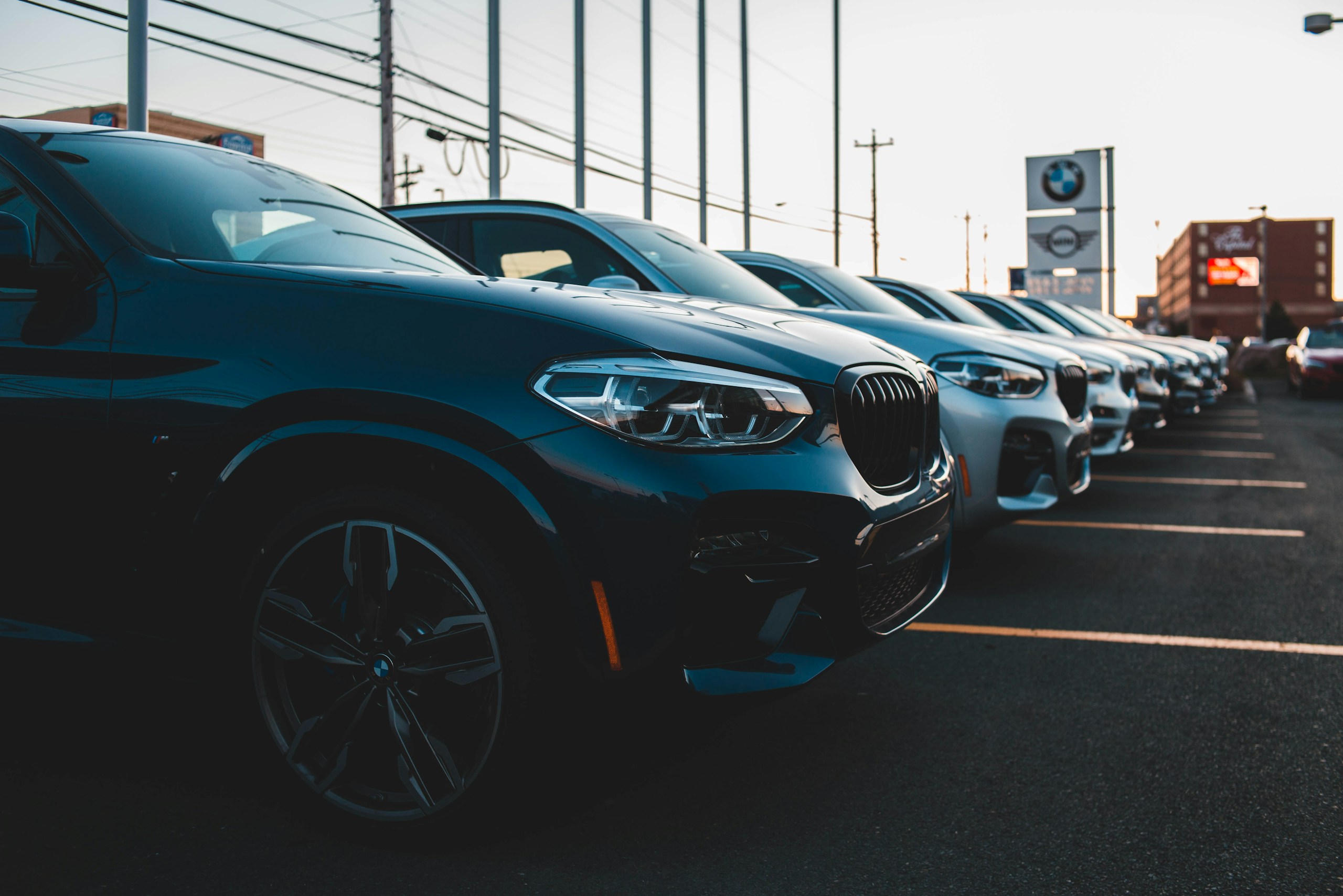 Row of BMW cars parked diagonally in a dealership lot, with signs and buildings in the background at dusk/sunset.
