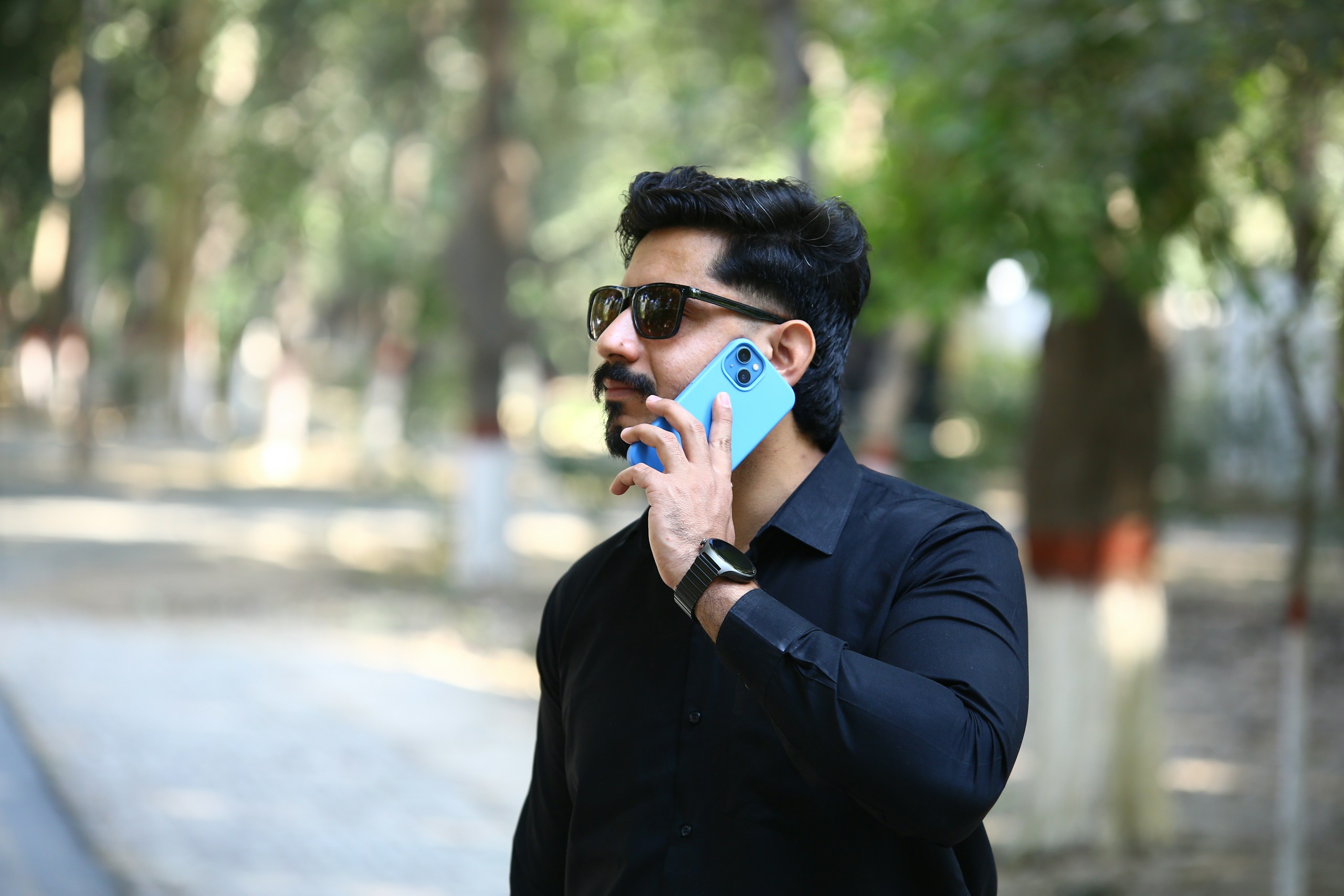 A stylish street-style portrait featuring black pants and shirt, sunglasses, and phone, captured at Kanan Park, Sahiwal. Perfect for wedding, event, or lifestyle-related content. Credit appreciated: ShadiCenter.com for providing this image.