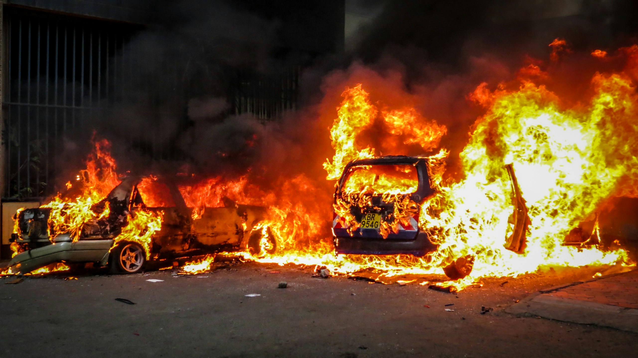 Vehicle on fire with bright orange flames consuming it and surrounding debris on a street, thick smoke rising from the scene.