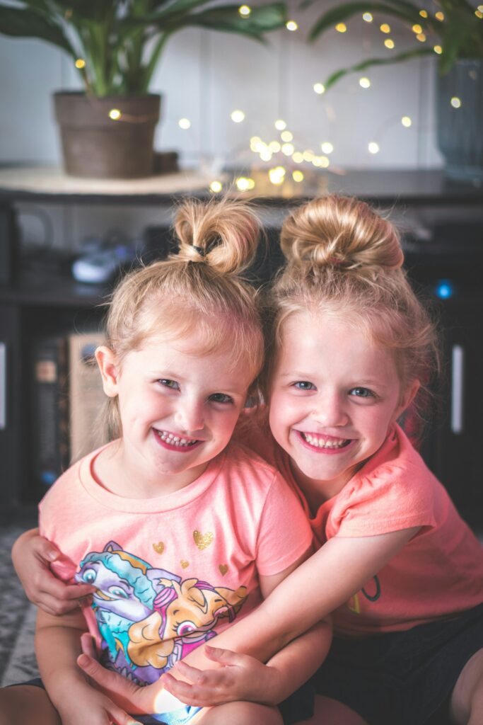 Two smiling young girls with their arms around each other, wearing pink tops, in a cozy room with blurred string lights in the background.