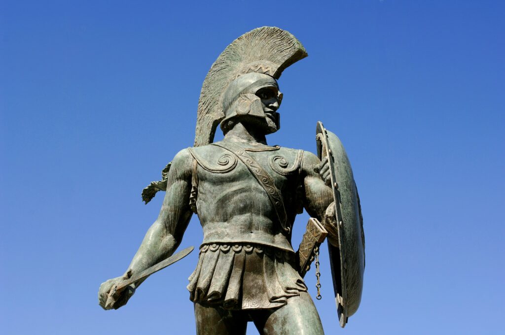 Bronze statue of a armored warrior with a tall crested helmet, gripping a sword in one hand and a shield in the other against a clear blue sky in the background, front-facing stance.
