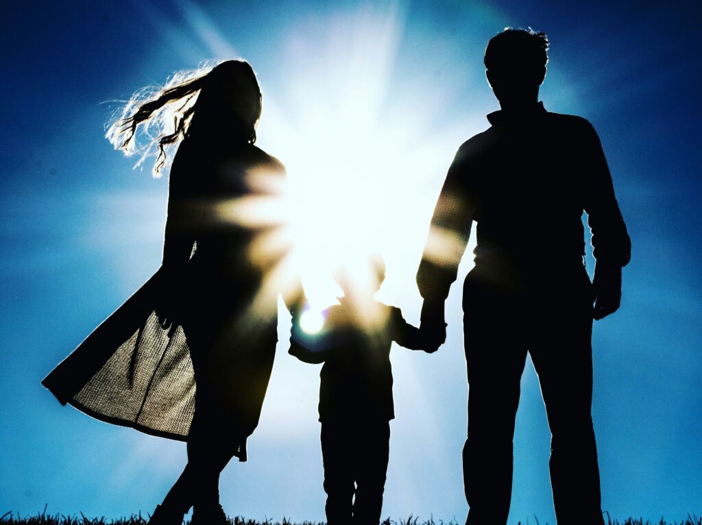 Silhouetted family of three holding hands outdoors, backlit by a bright sun with a blue sky.