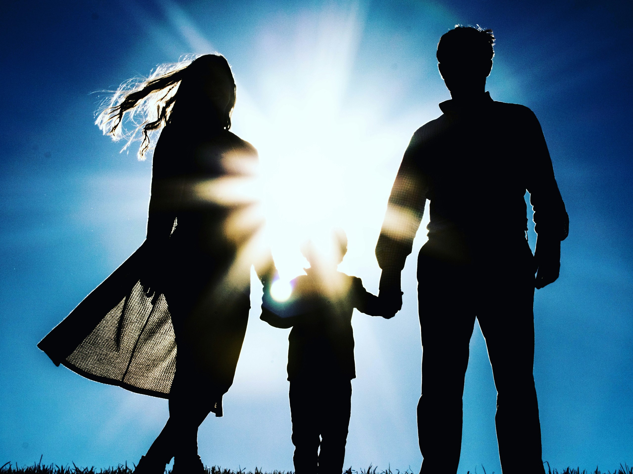 Silhouetted family of three holding hands outdoors, backlit by a bright sun with a blue sky.