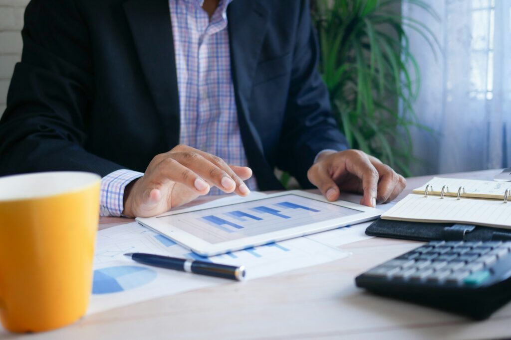 Businessperson in a dark blazer reviews charts on a tablet, with papers, a notebook, and a yellow mug on the desk and a plant in the background.