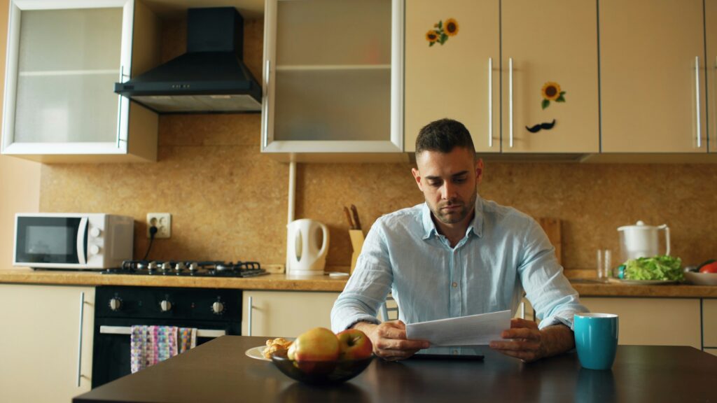 Upset young man reading letter with unpaid bill in the kitchen at home eraly morning