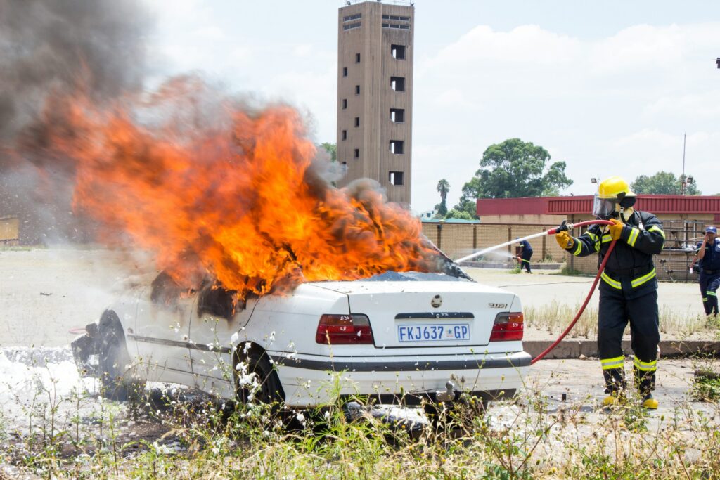 White sedan engulfed in bright orange flames with heavy black smoke; firefighter spraying water to extinguish the fire nearby at an outdoor lot.