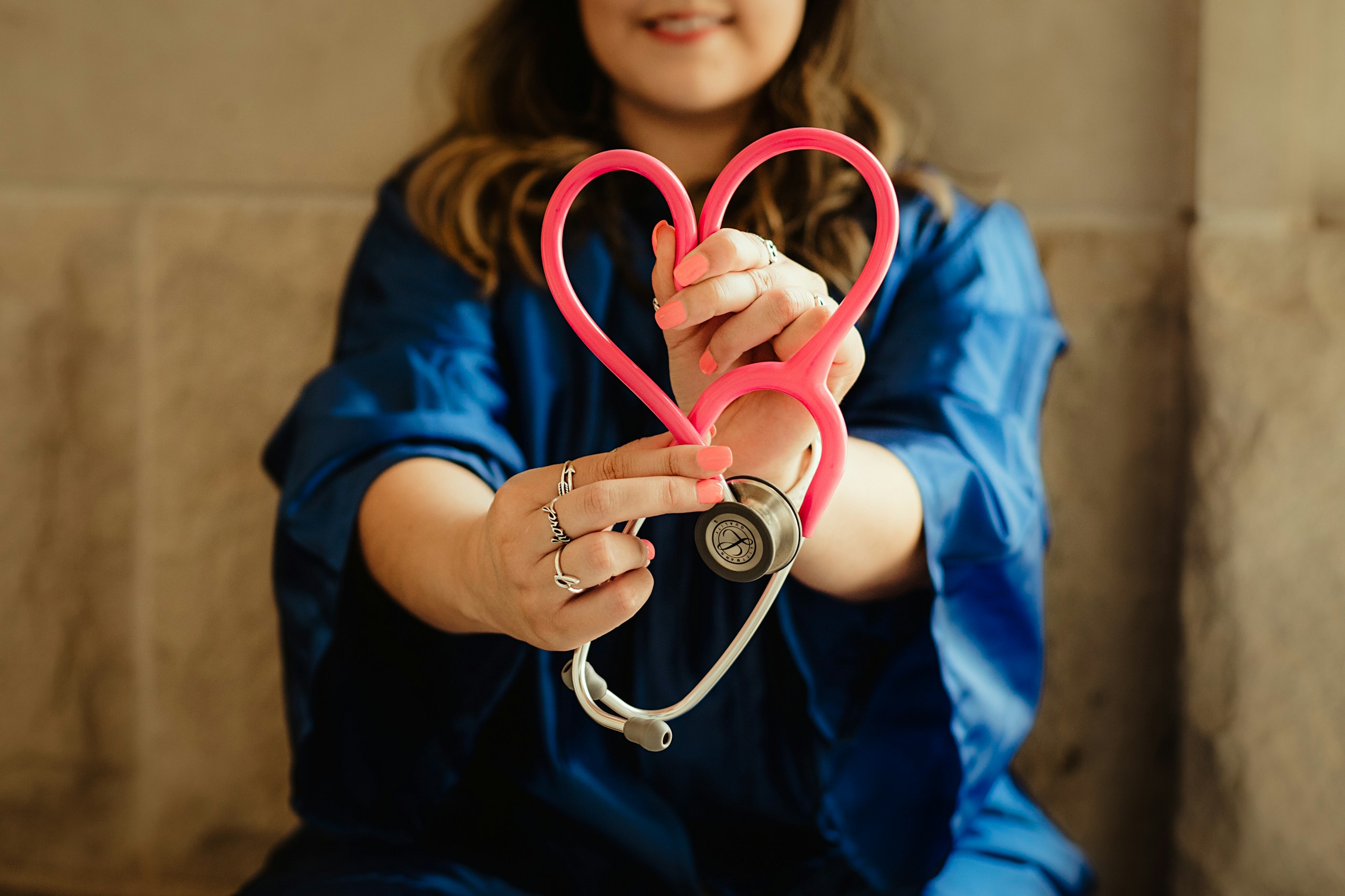 Person in blue scrubs holding a pink stethoscope shaped into a heart.
