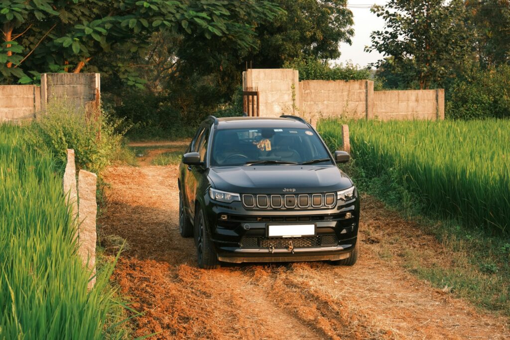 Black Jeep SUV parked on a dirt path between tall grasses and a low concrete wall.