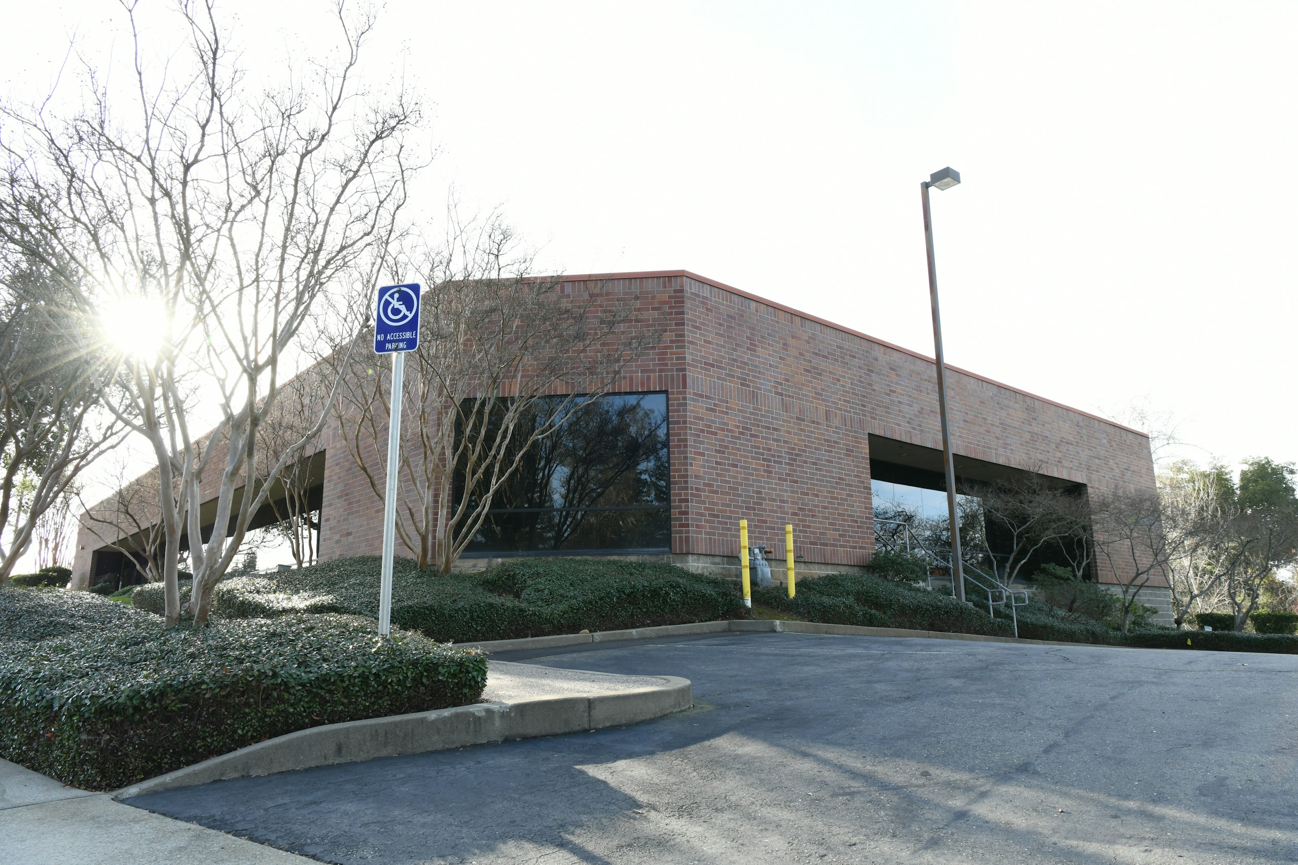 Blue handicap parking sign reading 'No Accessible Parking' in front of a brick office building with leafless trees and a sun flare on the left side of the image.