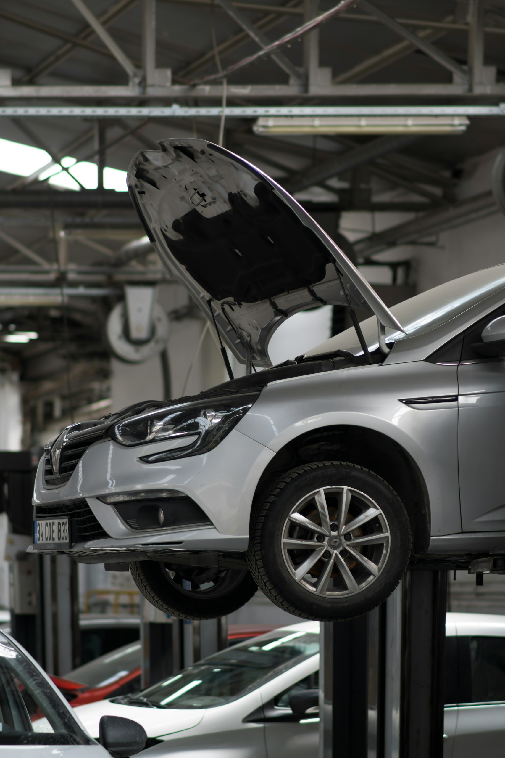 Silver SUV raised on a car lift with its hood open in an auto repair shop, industrial ceiling and equipment visible in background