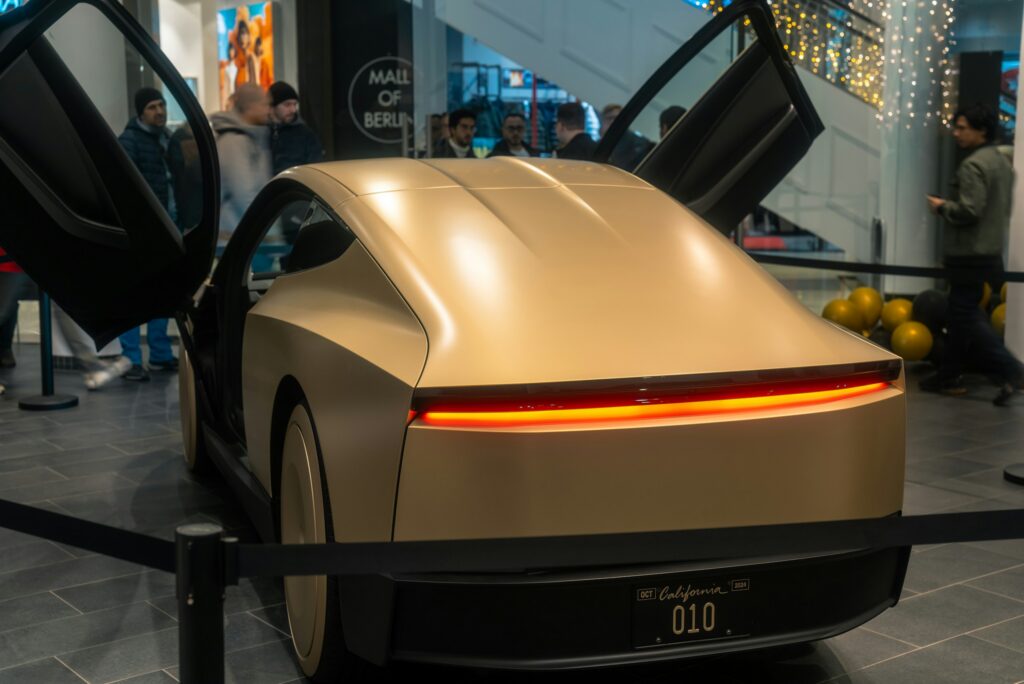 Gold sports car with scissor doors open on display in a showroom; crowd in the background and a California license plate reading 010