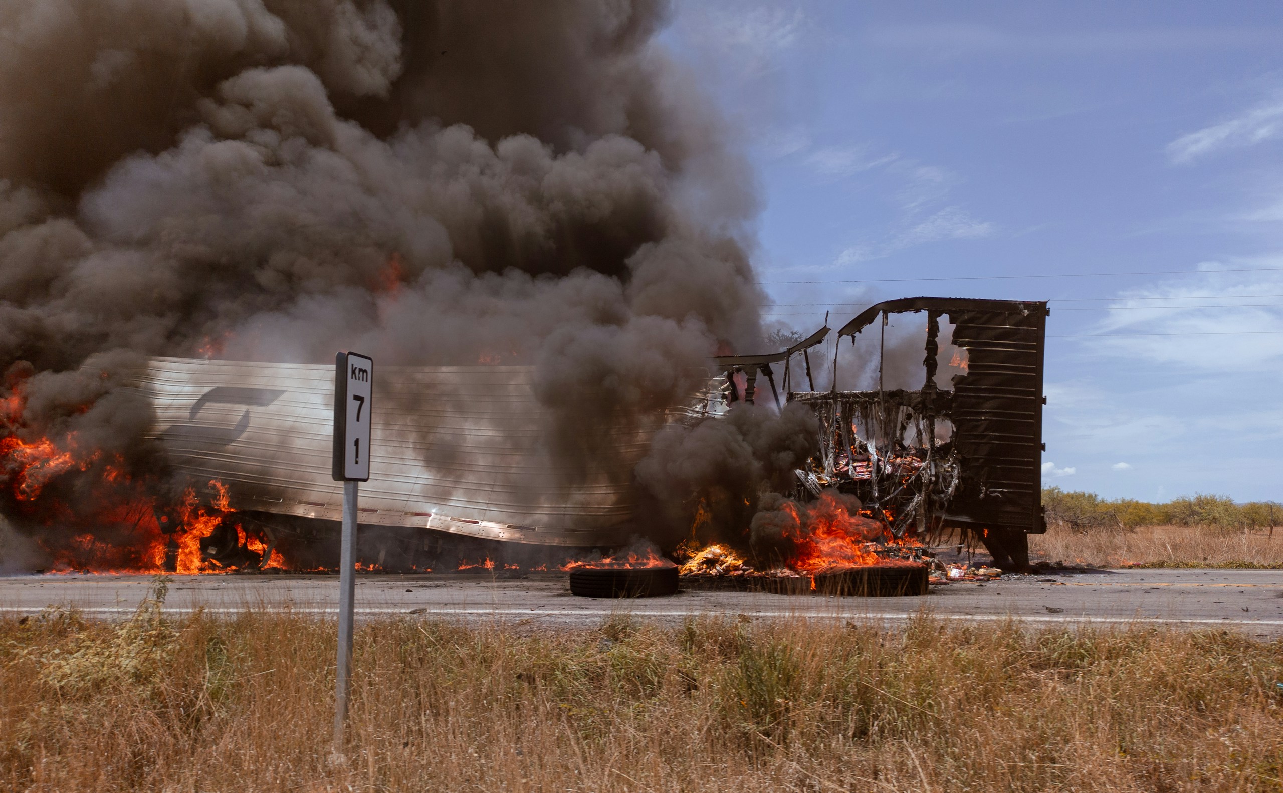 A burning railcar fire with thick black smoke billowing upward, flames at the base, near a roadside sign that reads 'km 7' in the foreground.