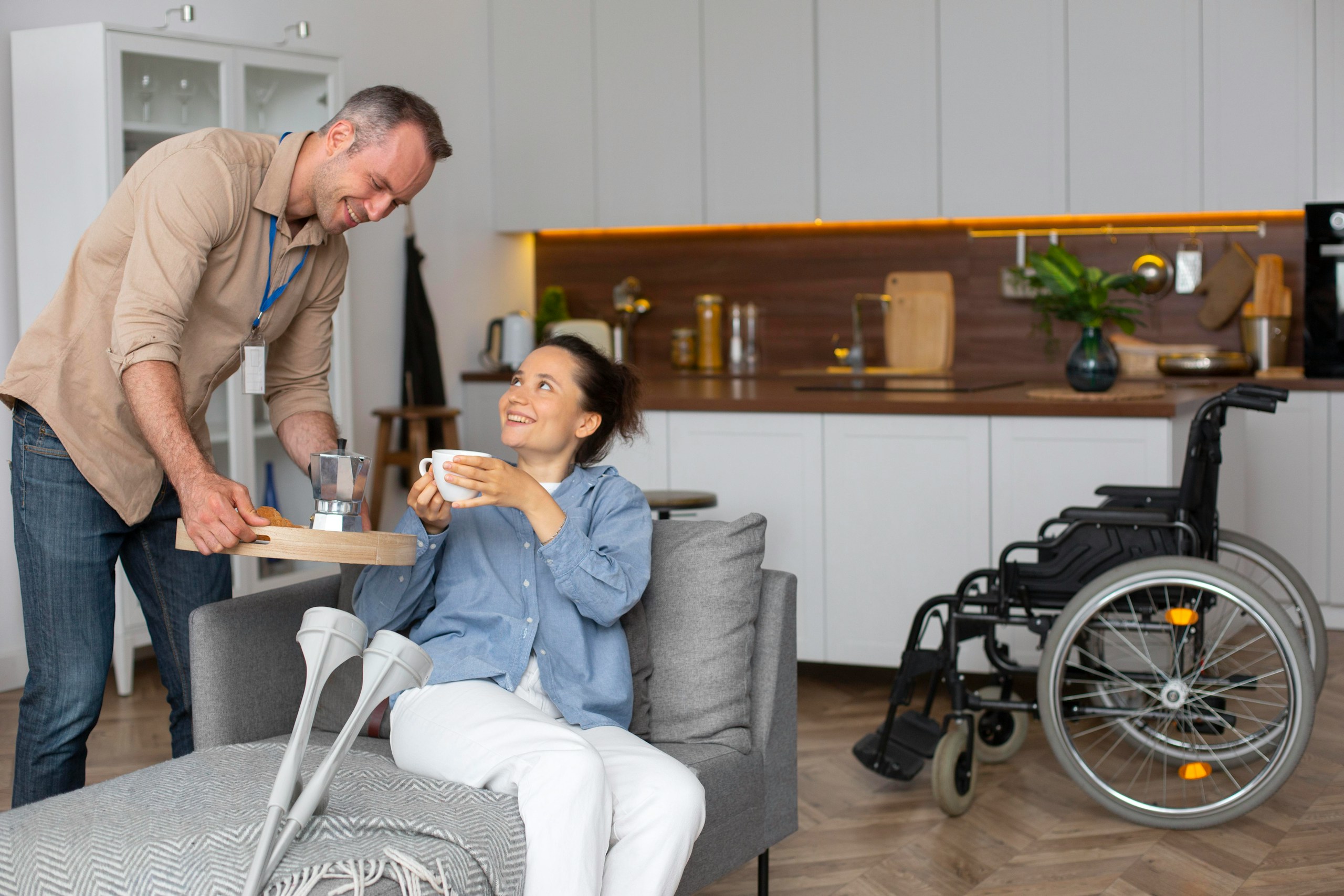 Man standing and smiling as he serves a tray of cookies to a woman in a wheelchair in a bright, modern kitchen.
