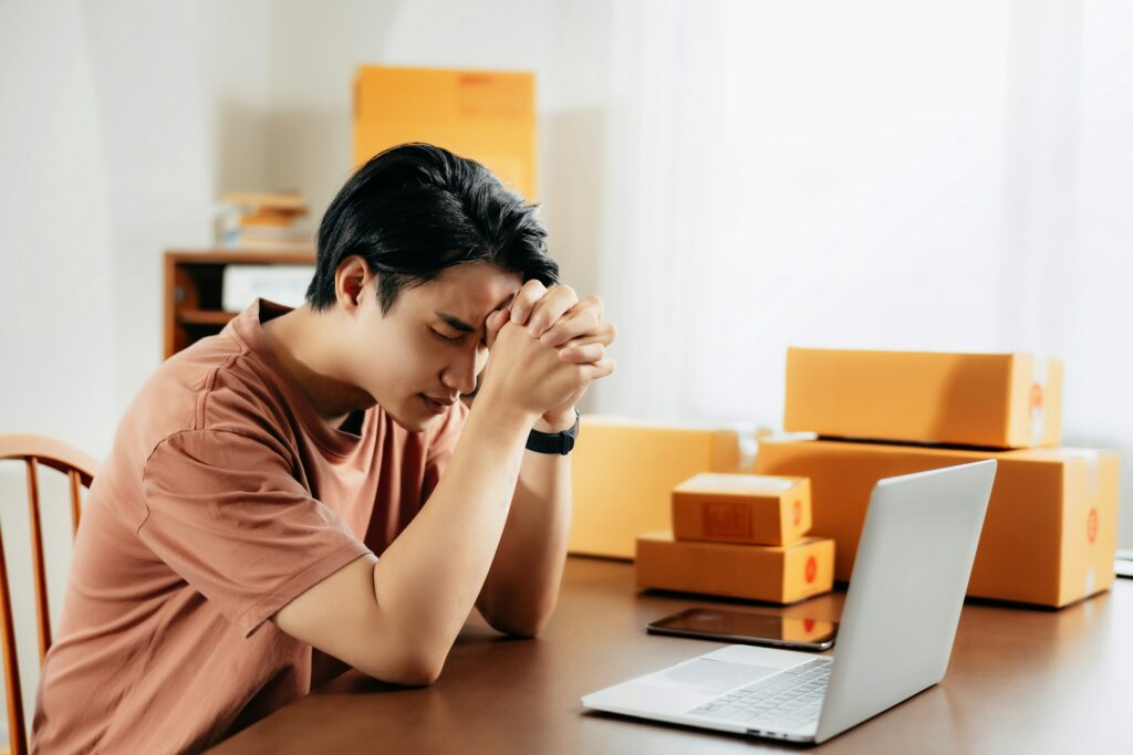 Young man sits at a table with hands clasped over his forehead, looking stressed amid stacked orange boxes and a laptop in a home office.