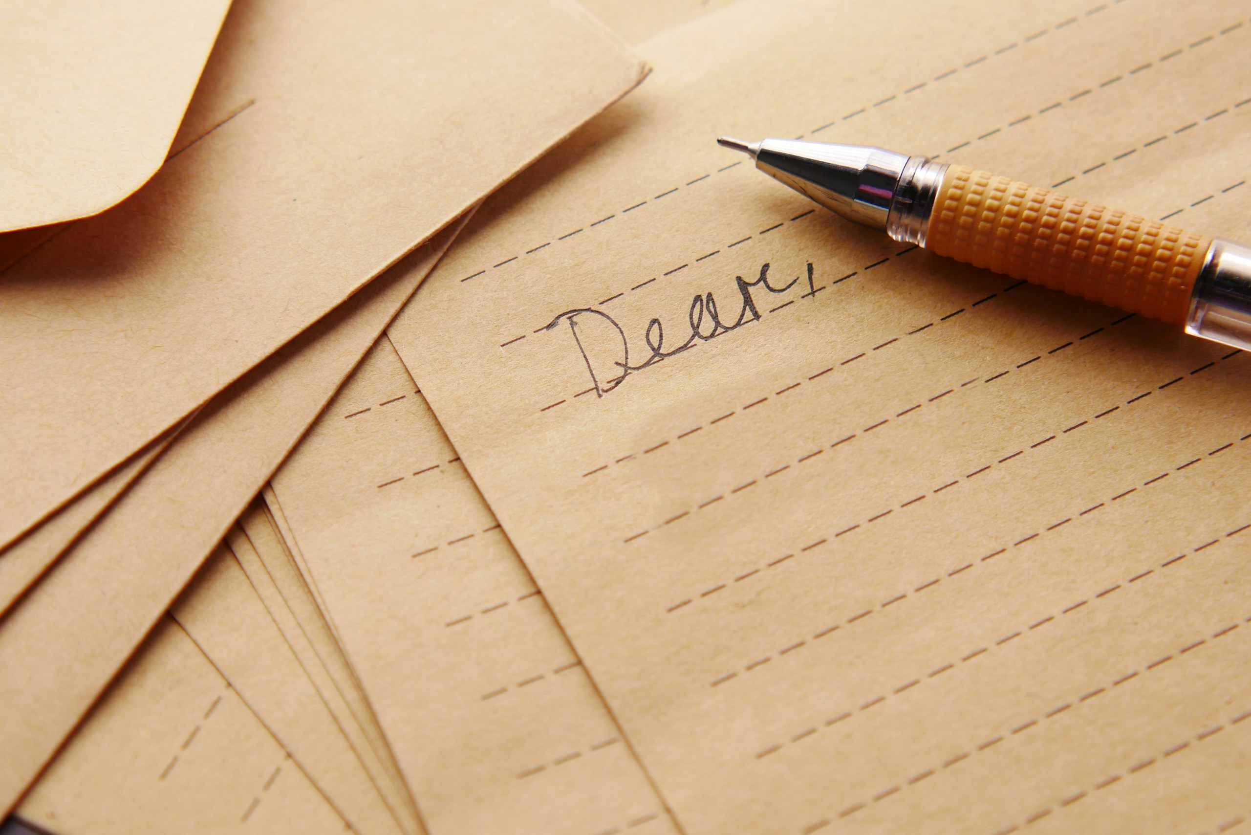 Close-up of a fountain pen nib resting on tan lined paper with the handwritten word 'Dear' in cursive.