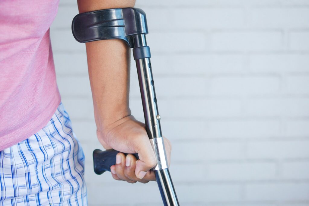 Person gripping a blue-and-silver forearm crutch, wearing a pink shirt and blue plaid shorts, against a pale brick wall.
