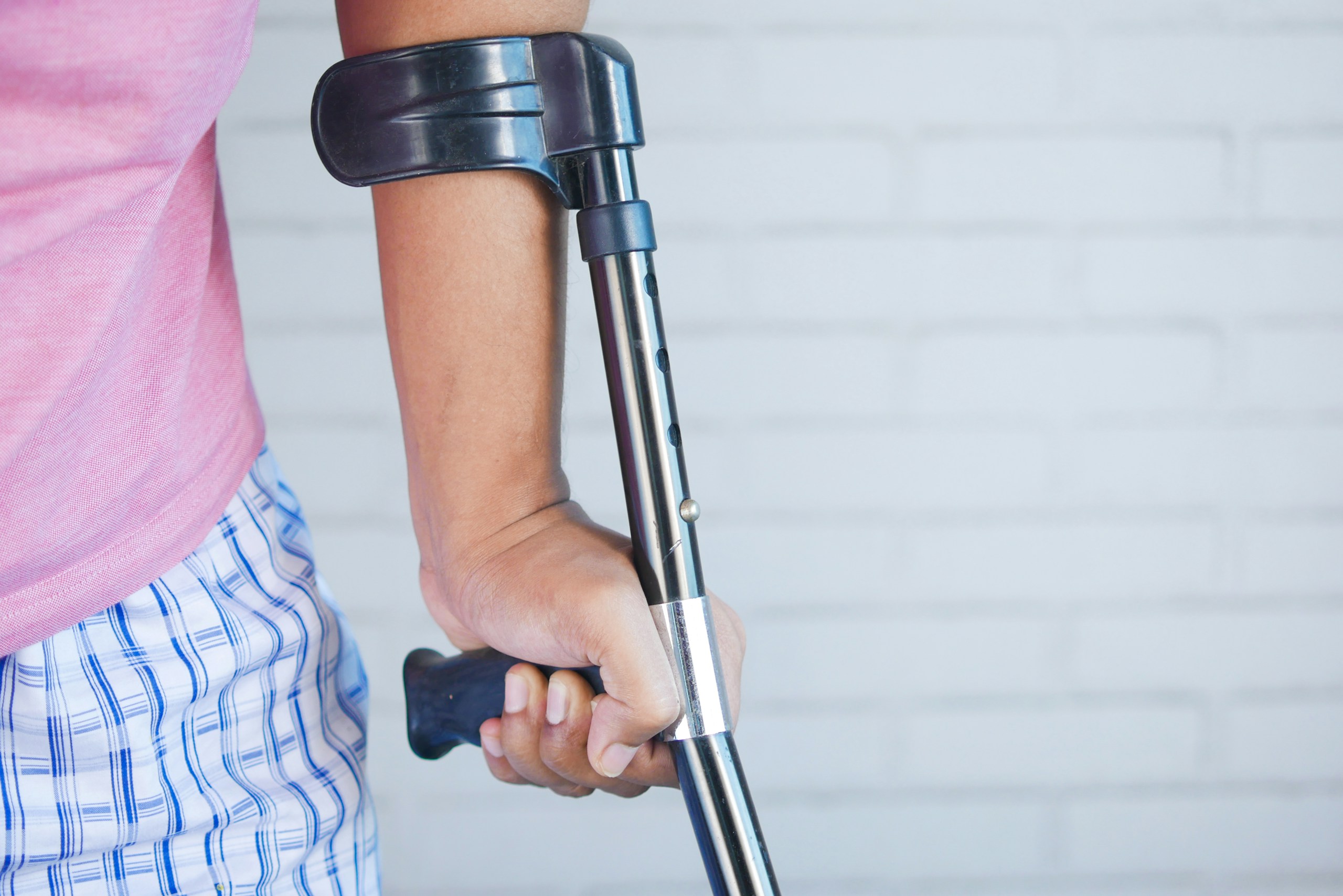 Person gripping a blue-and-silver forearm crutch, wearing a pink shirt and blue plaid shorts, against a pale brick wall.