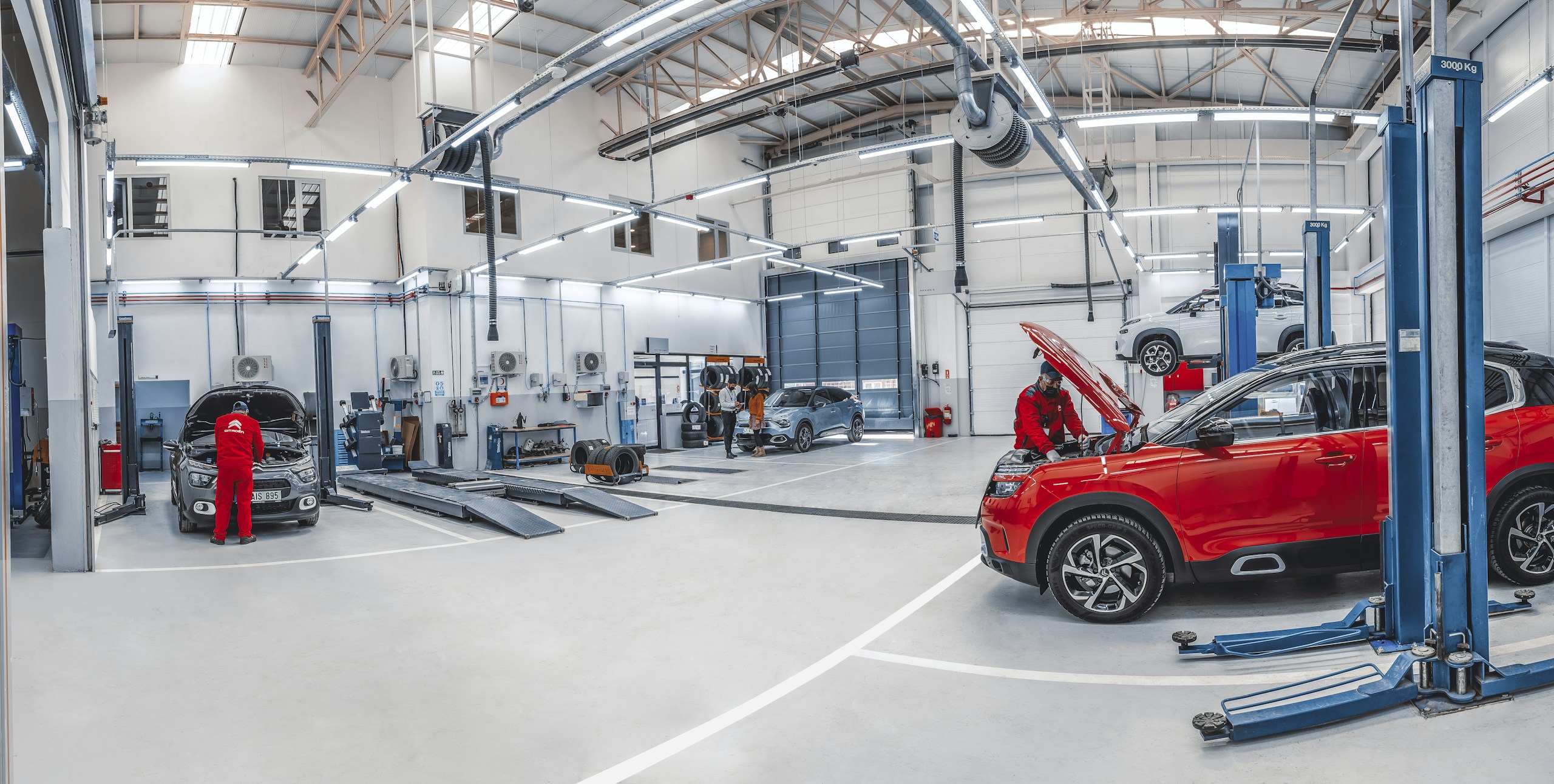 Wide view of a modern auto repair shop with a red SUV in the foreground and technicians in red uniforms working on cars.