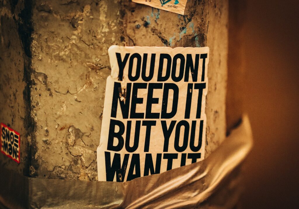 Close-up of a worn wall with a bold poster shouting: 'YOU DONT NEED IT BUT YOU WANT IT' against a textured, yellowed surface.