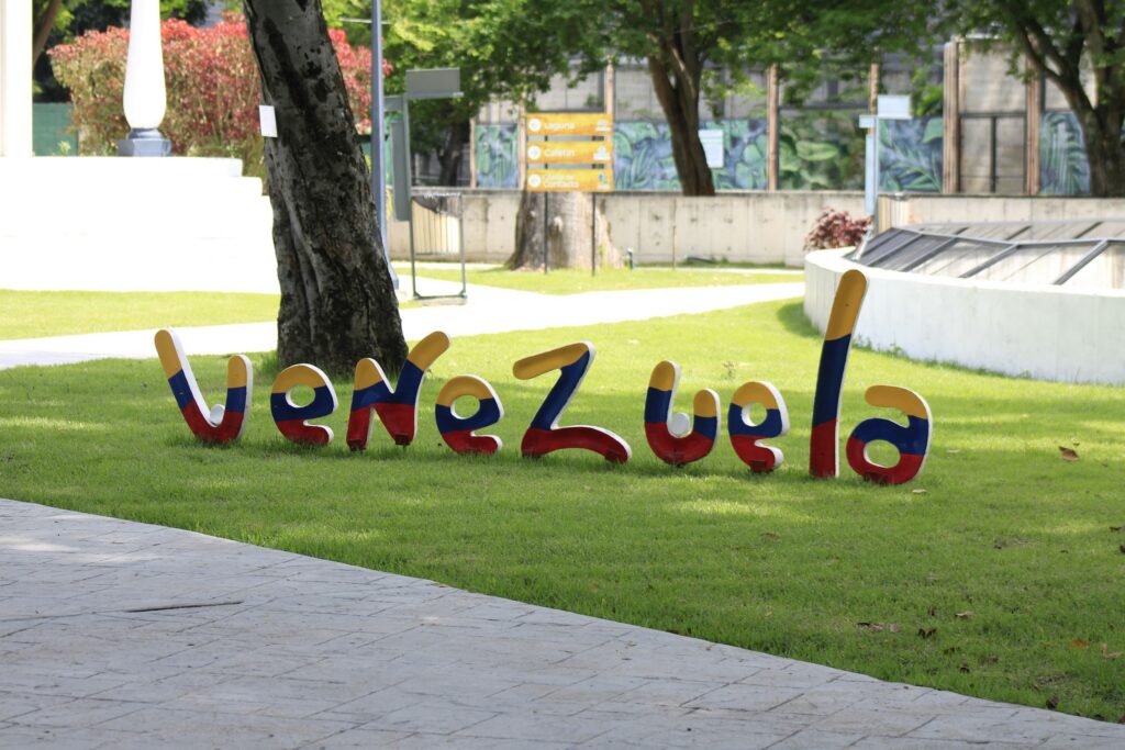 Colorful three-dimensional letters spelling 'Bienvenidos' arranged on the green lawn in a park setting with trees in the background.