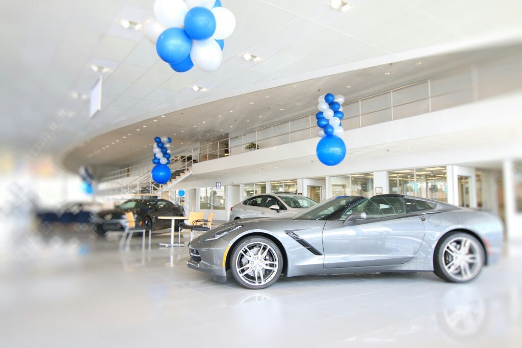 New car showroom with a silver sports car in the foreground and blue-and-white balloon decorations hanging from the ceiling and columns.