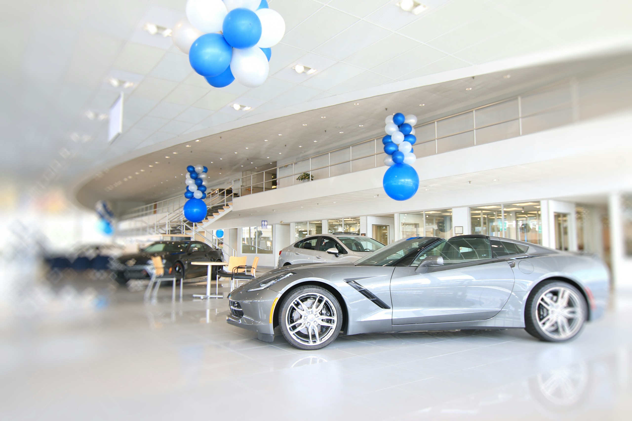 New car showroom with a silver sports car in the foreground and blue-and-white balloon decorations hanging from the ceiling and columns.