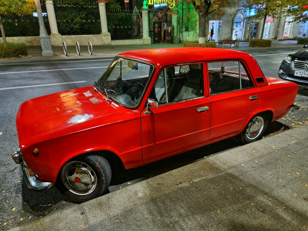 Red vintage sedan parked along a city sidewalk, storefronts and trees visible in the background.