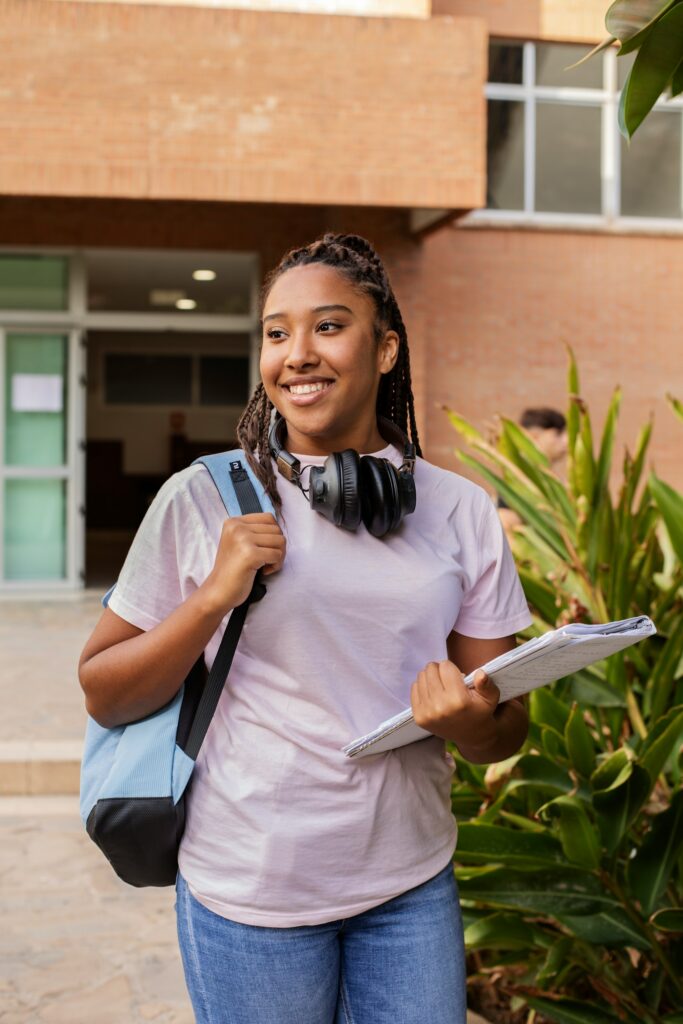 Young woman with braids, wearing a light pink T‑shirt and jeans, carries a blue backpack, headphones, and papers outside a brick building.