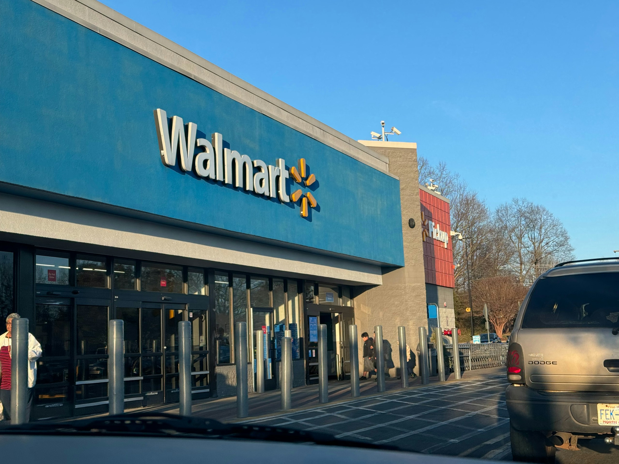 Front view of a Walmart store with a blue façade and large white Walmart lettering; a grey SUV is parked at the curb in the foreground.
