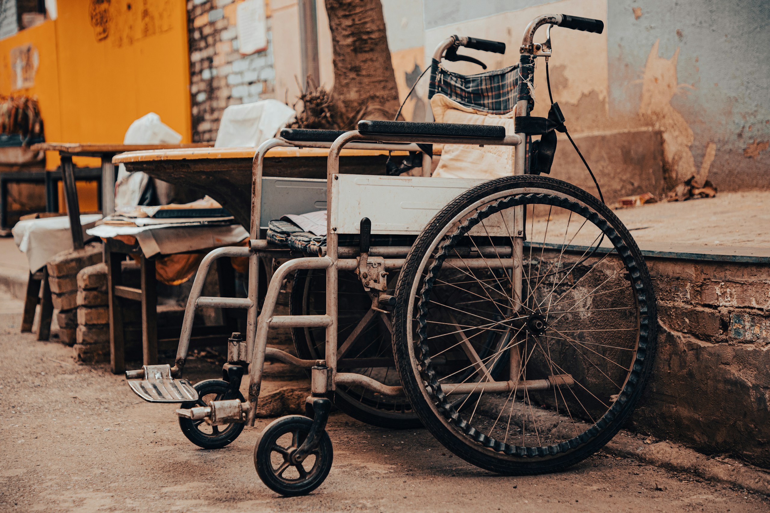 Wheelchair attached to a flatbed cart in a gritty alley with peeling walls and orange tones in the background.