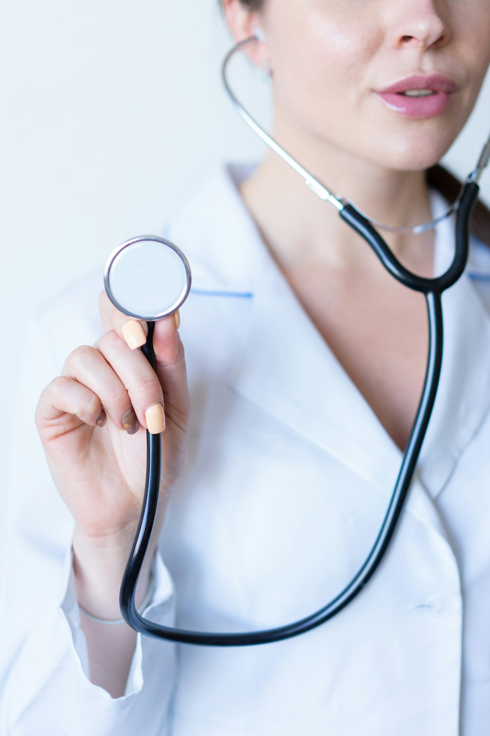 Medical professional holding a stethoscope toward the camera with a white lab coat visible at the edges.