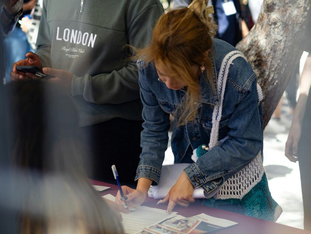 People gathered around a table signing or filling out documents, with a person in a denim jacket nearby.