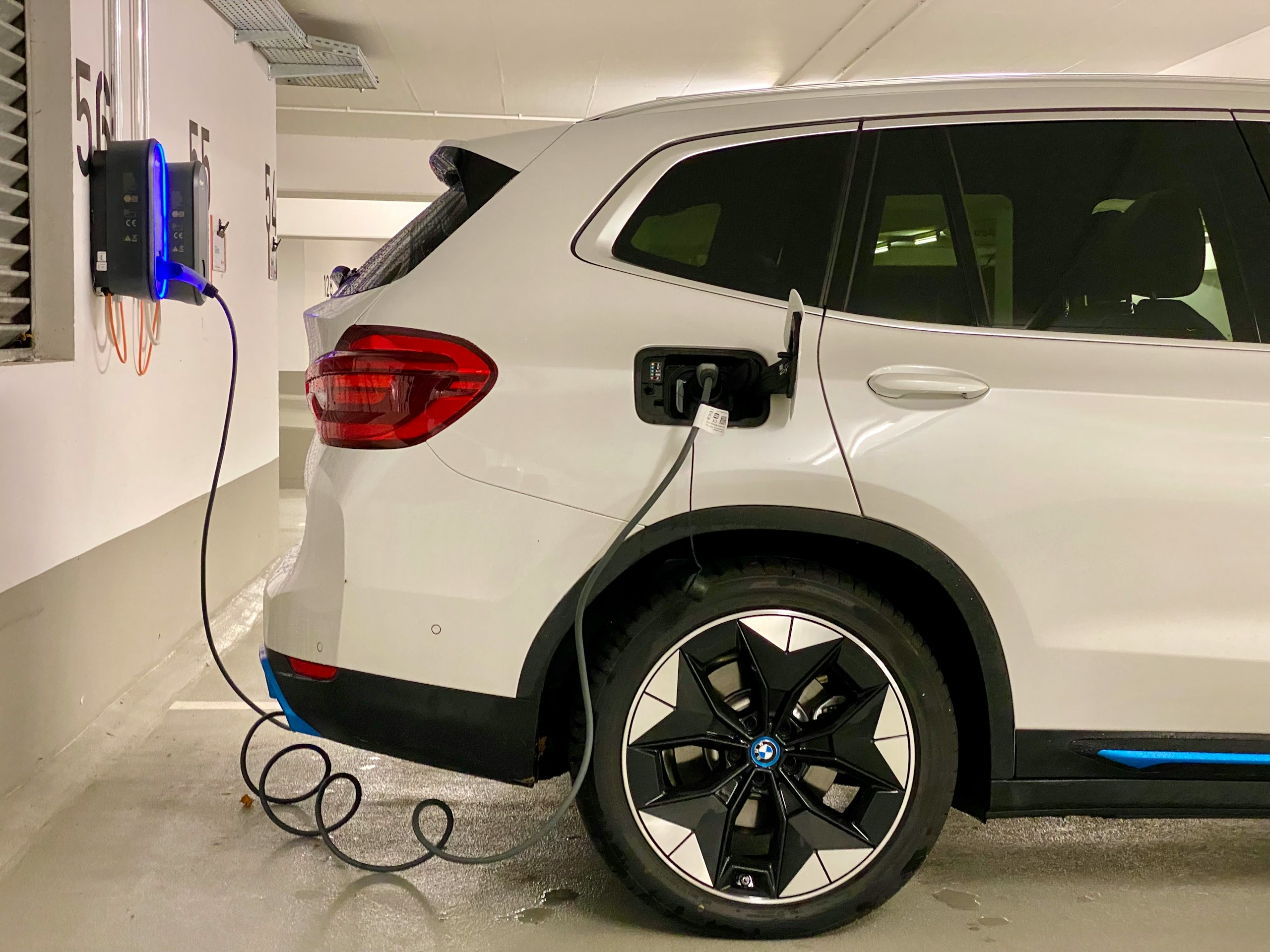 White BMW SUV being charged at a wall-mounted EV charger in a parking garage, blue charging cable connected to the rear left side.