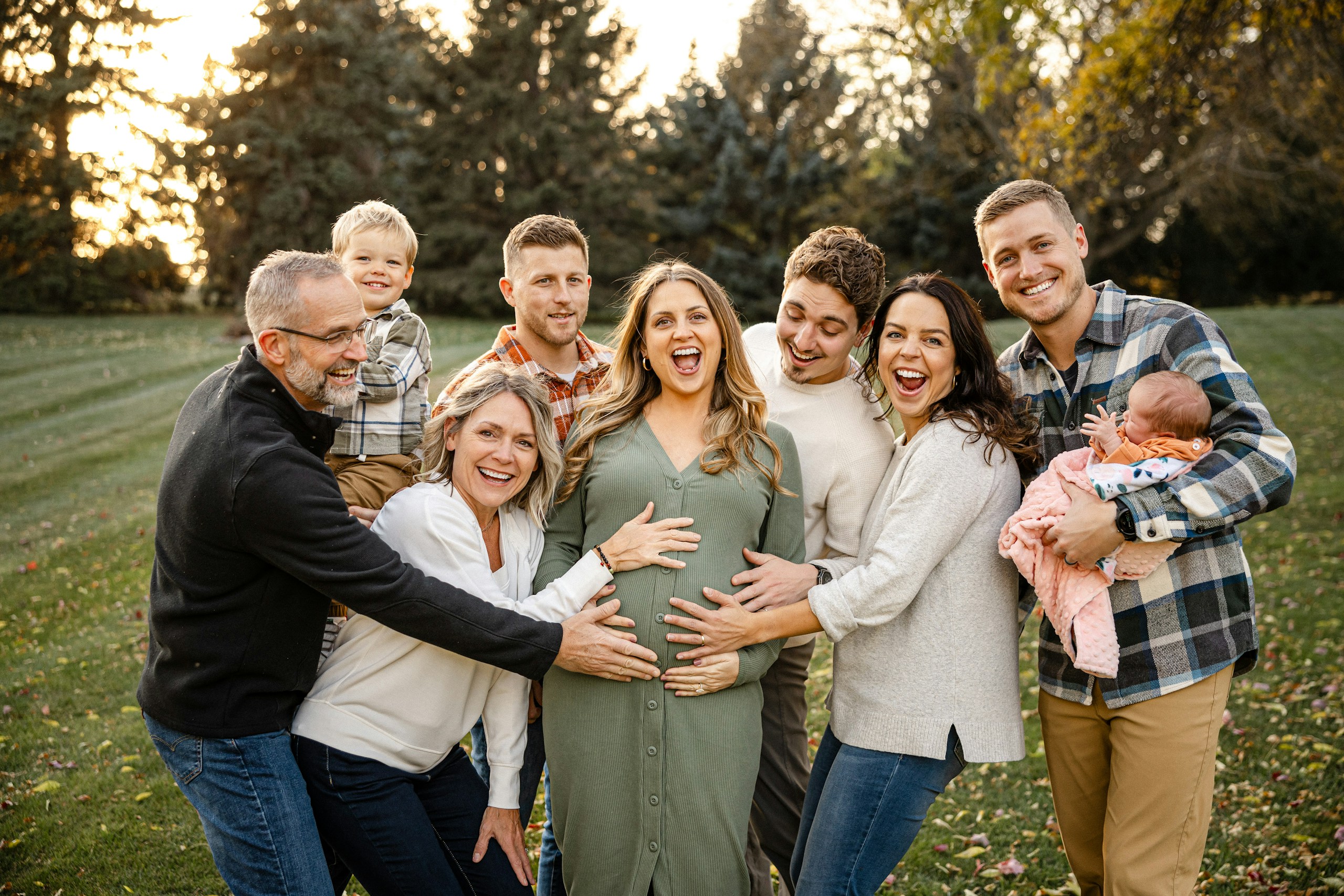 Group of smiling adults outdoors with a pregnant woman at center, posing as a family photo in a park.