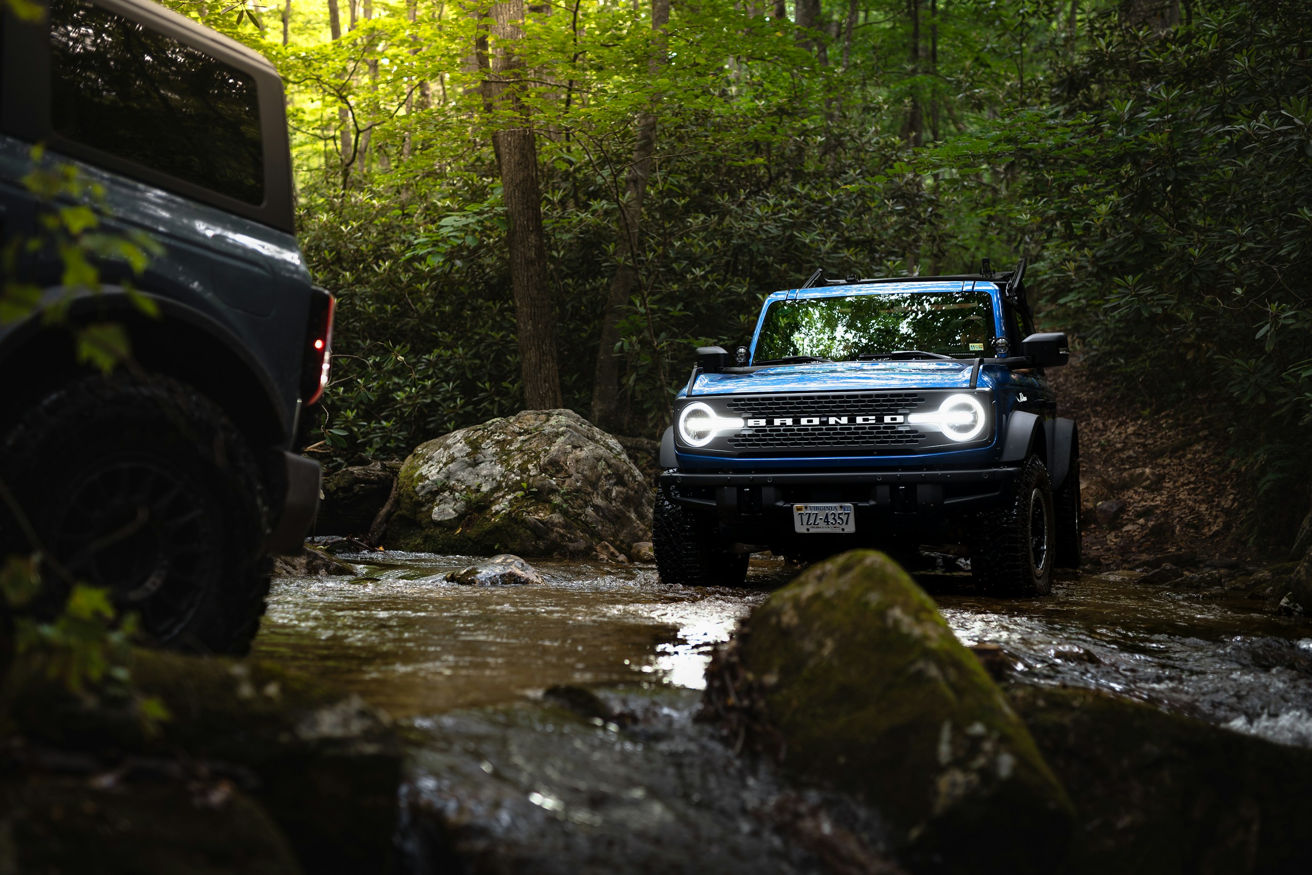 Blue Ford Bronco off-roader driving through a rocky forest stream, with another vehicle blurred on the left.