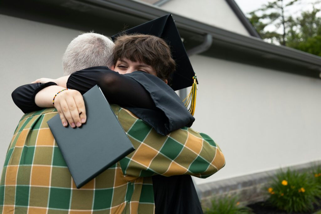 Graduate in cap and gown hugs an older person outdoors, holding a diploma.