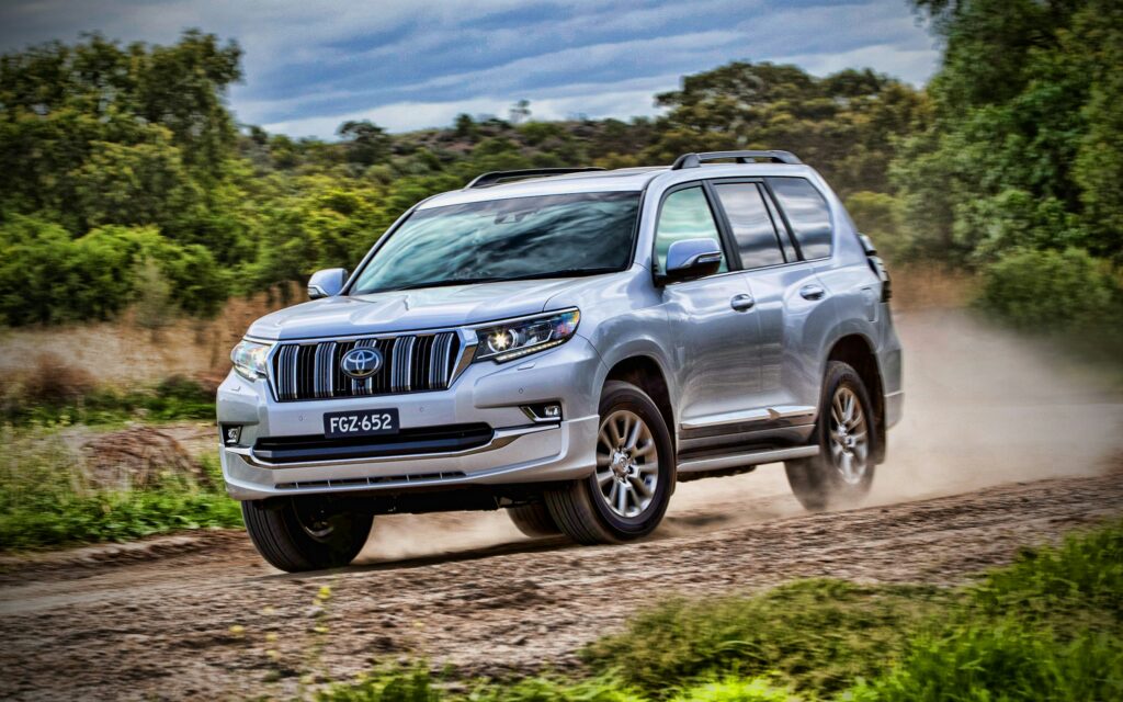 Silver SUV driving on a dirt road through grassy countryside, kicking up dust behind it.