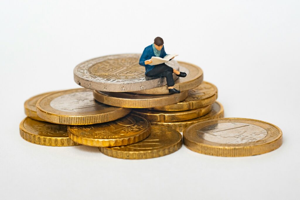 Small figurine of a man in a blue jacket sitting on a stack of coins, reading a newspaper, symbolizing financial news or wealth planning